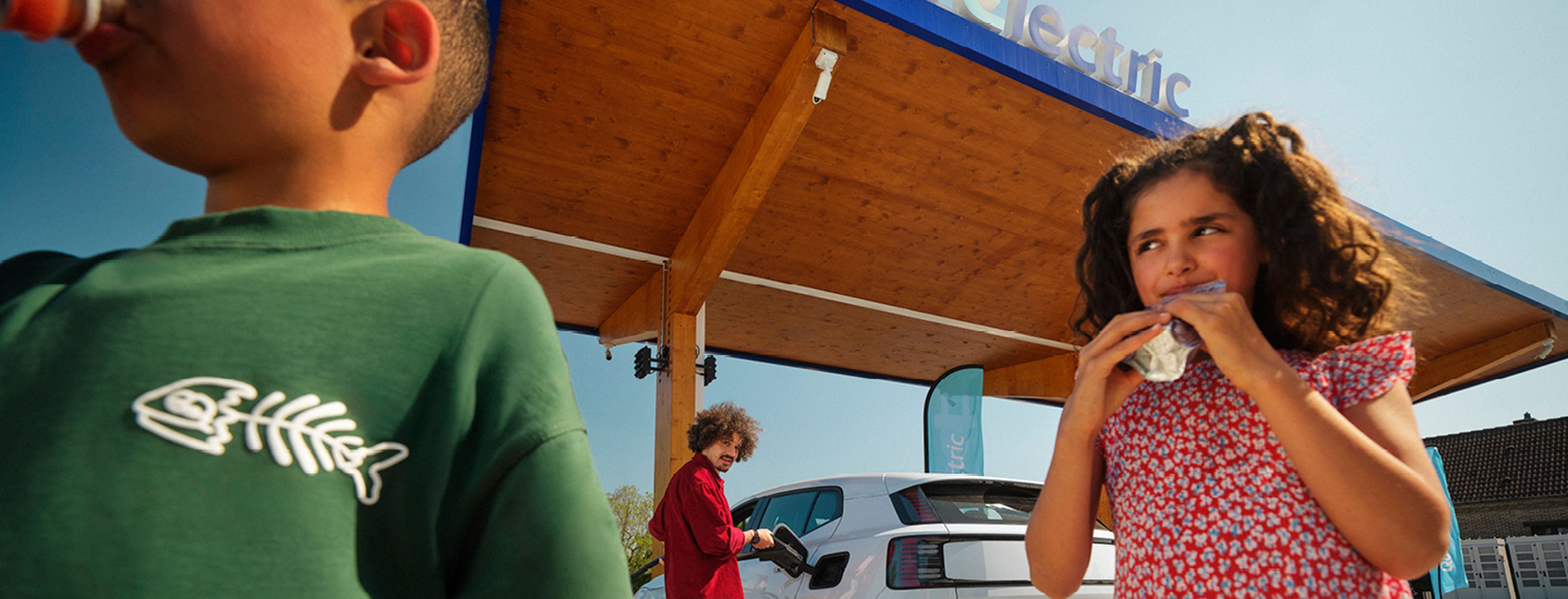 Enfants boivent pendant qu'un homme recharge une voiture électrique à une station Q8 Electric