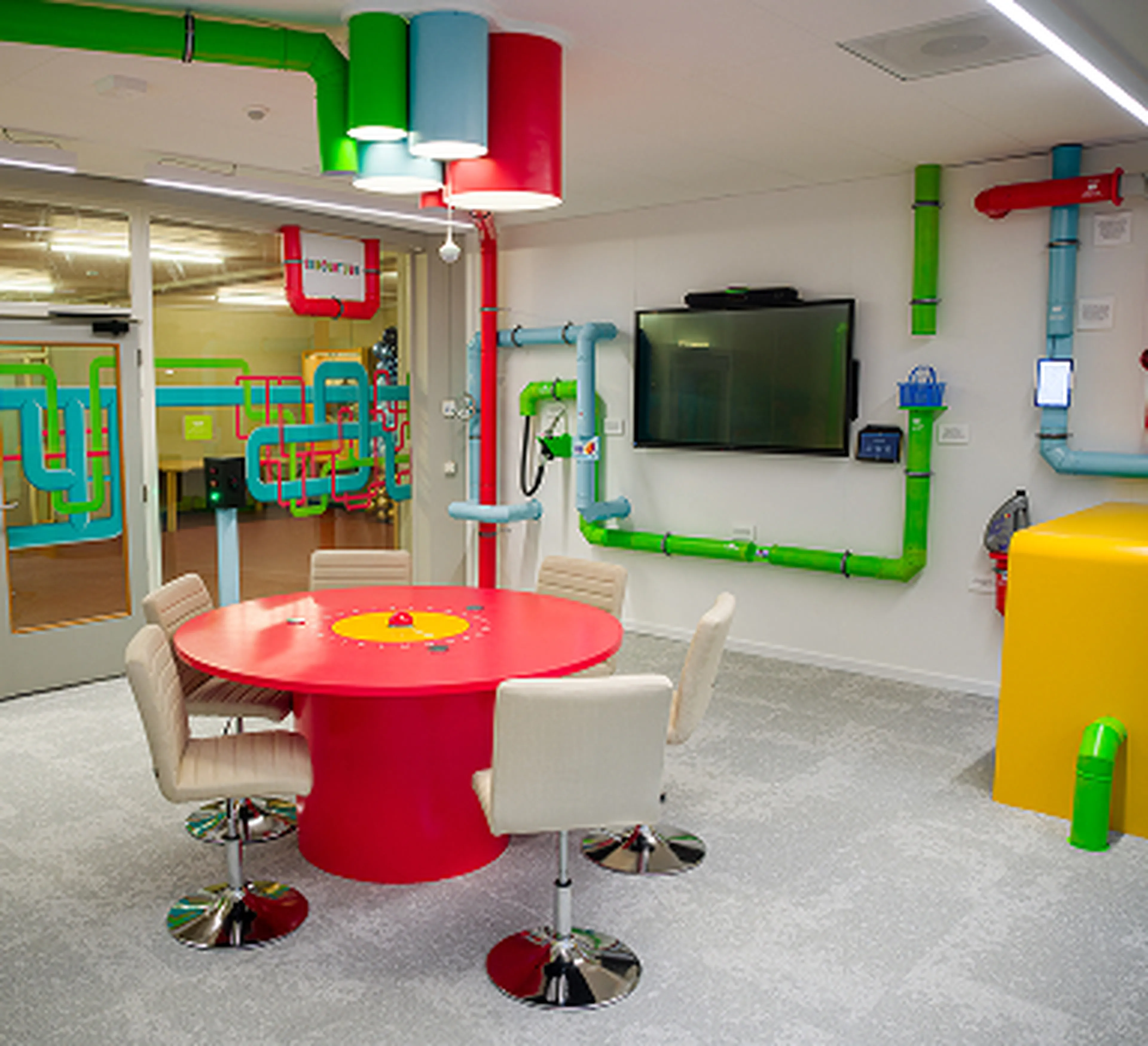 Salle de réunion au design ludique au siège de Q8 à Rotterdam, avec une table ronde rouge vif, des chaises blanches et des tuyaux colorés au plafond et sur les murs. Un écran de télévision est fixé au centre du mur.