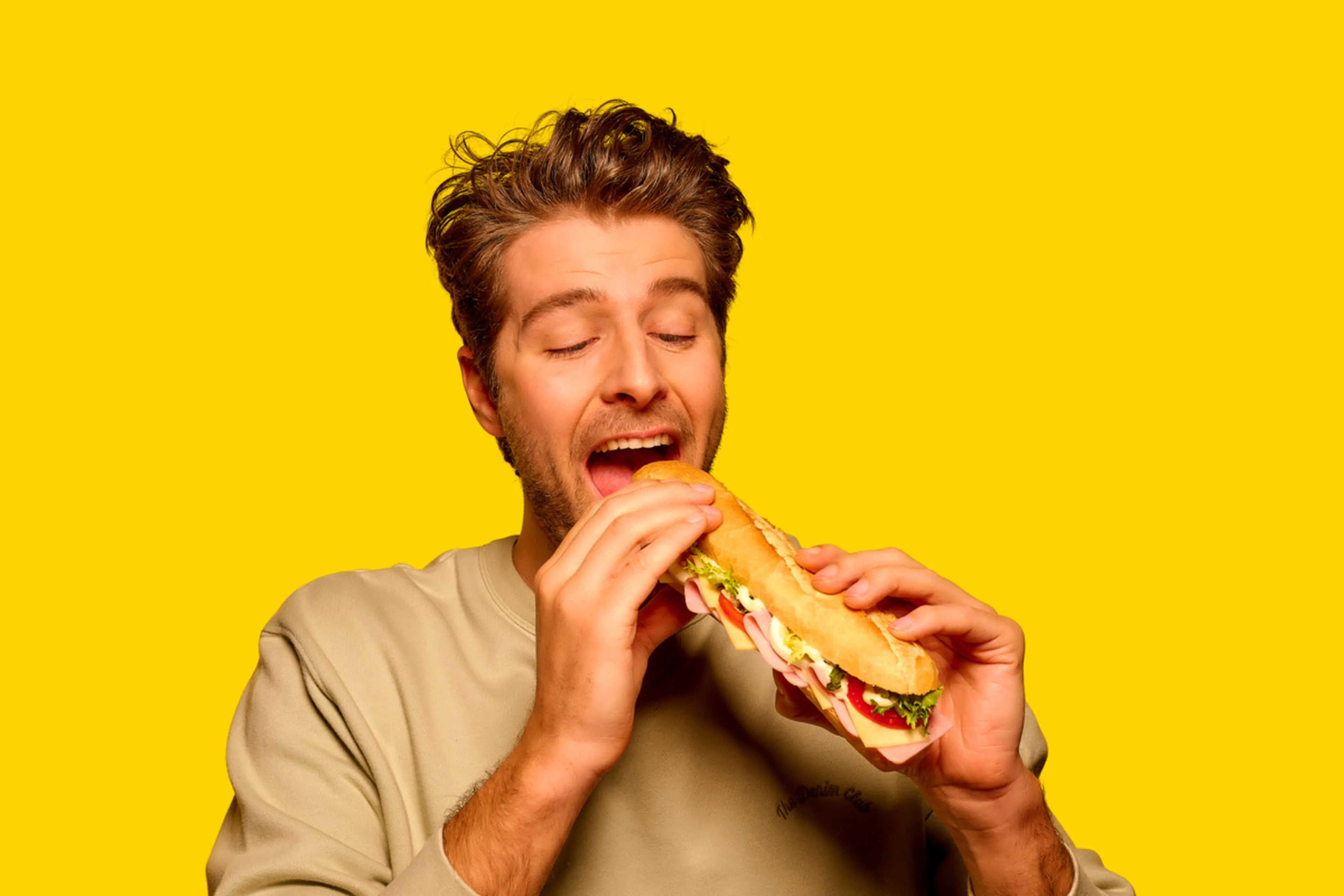 Un homme aux cheveux châtain clair, portant un pull beige, se tient devant un fond jaune vif et prend une grande bouchée d’un sandwich garni, les yeux fermés.