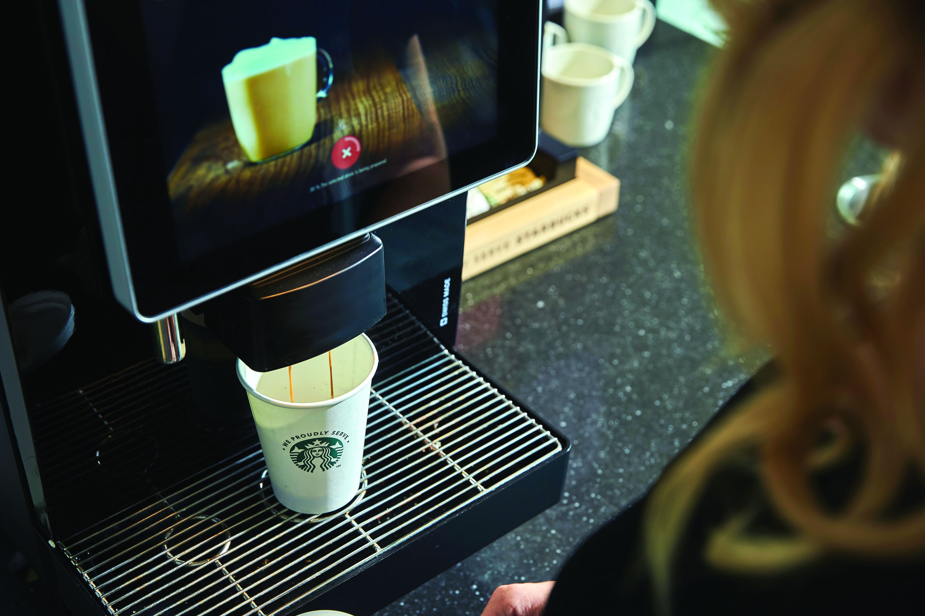 Machine à café Starbucks préparant une boisson chaude dans un gobelet, avec écran tactile et une personne à l’arrière-plan dans une station Q8.