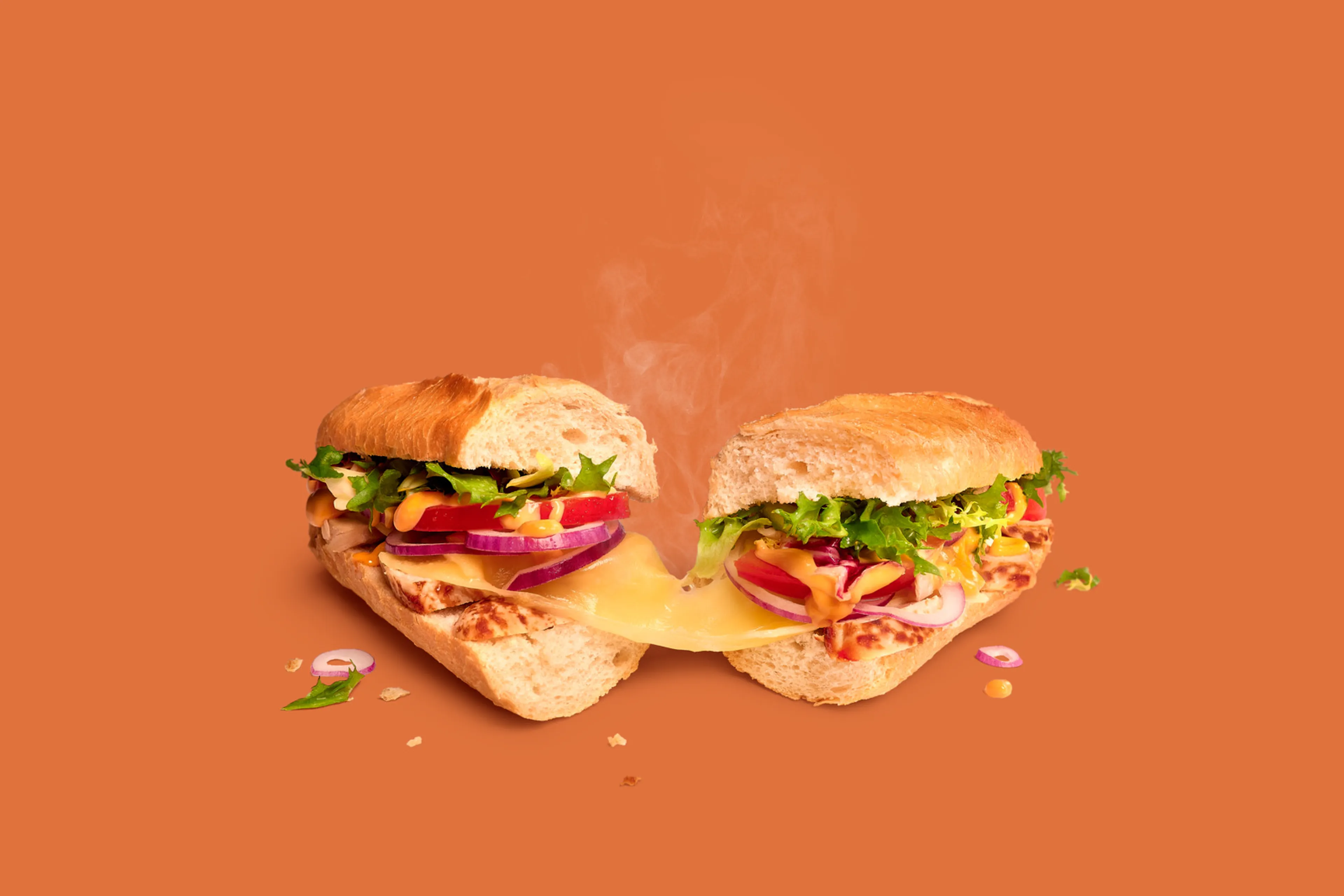 Sandwich Chicken Samouraï Remix de Panos, coupé en deux, avec poulet, fromage, légumes et sauce sur fond orange.