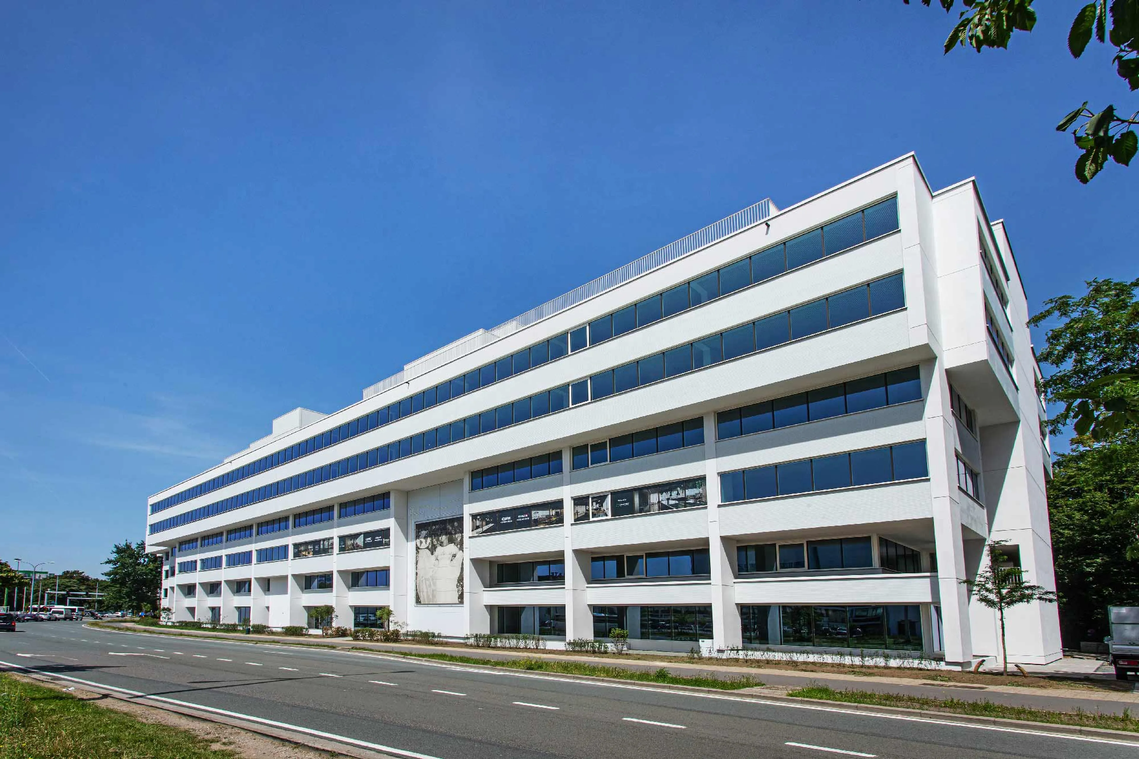 Siège social de Q8 à Anvers, faisant partie de la Greenhouse Collection — un bâtiment de bureaux moderne et blanc, avec des fenêtres horizontales sur plusieurs étages, situé le long d’une large avenue urbaine sous un ciel bleu clair.