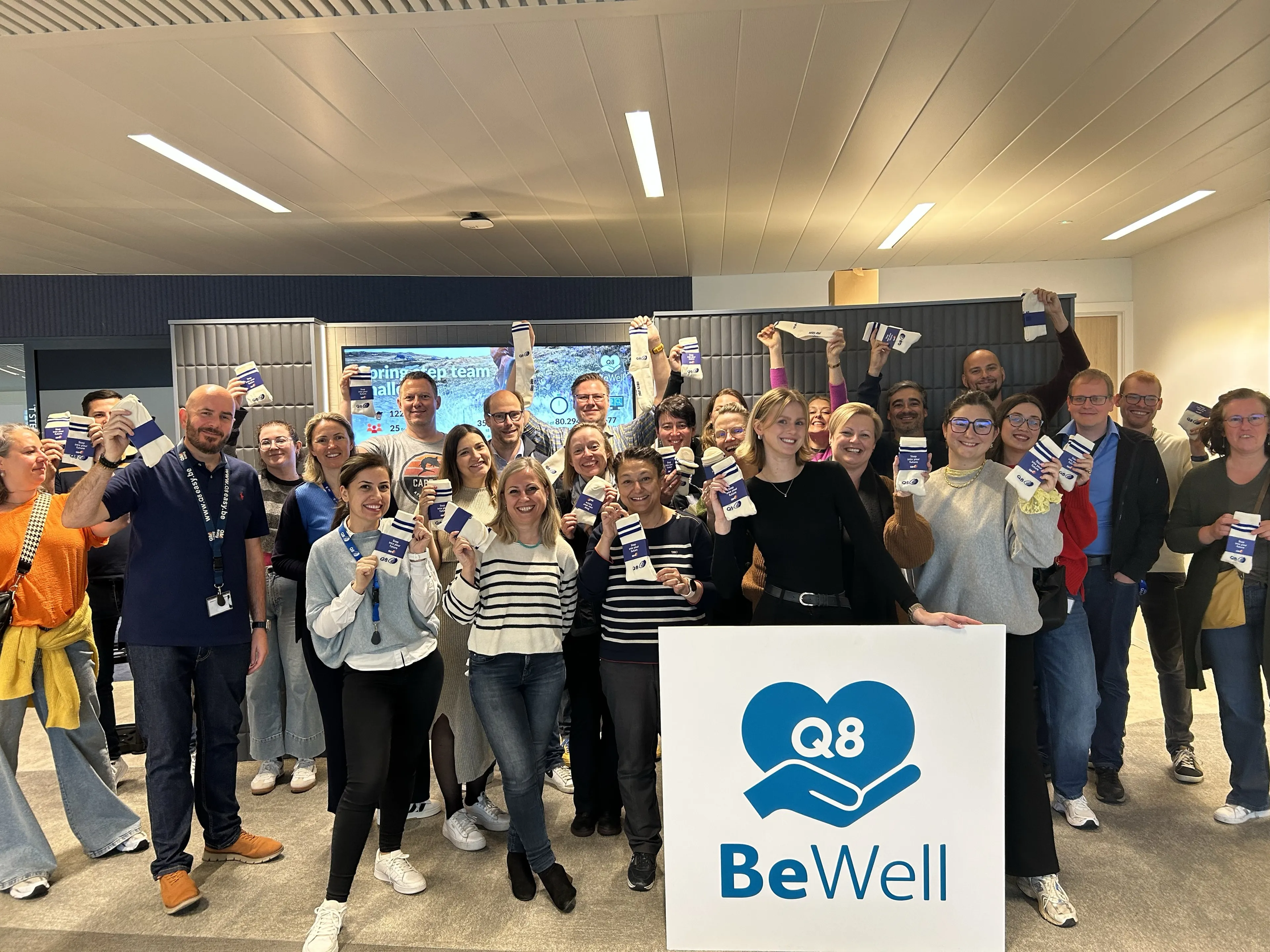 Un grand groupe de collègues Q8 sourit à la caméra et prend une photo de groupe avec une pancarte BeWell devant eux.