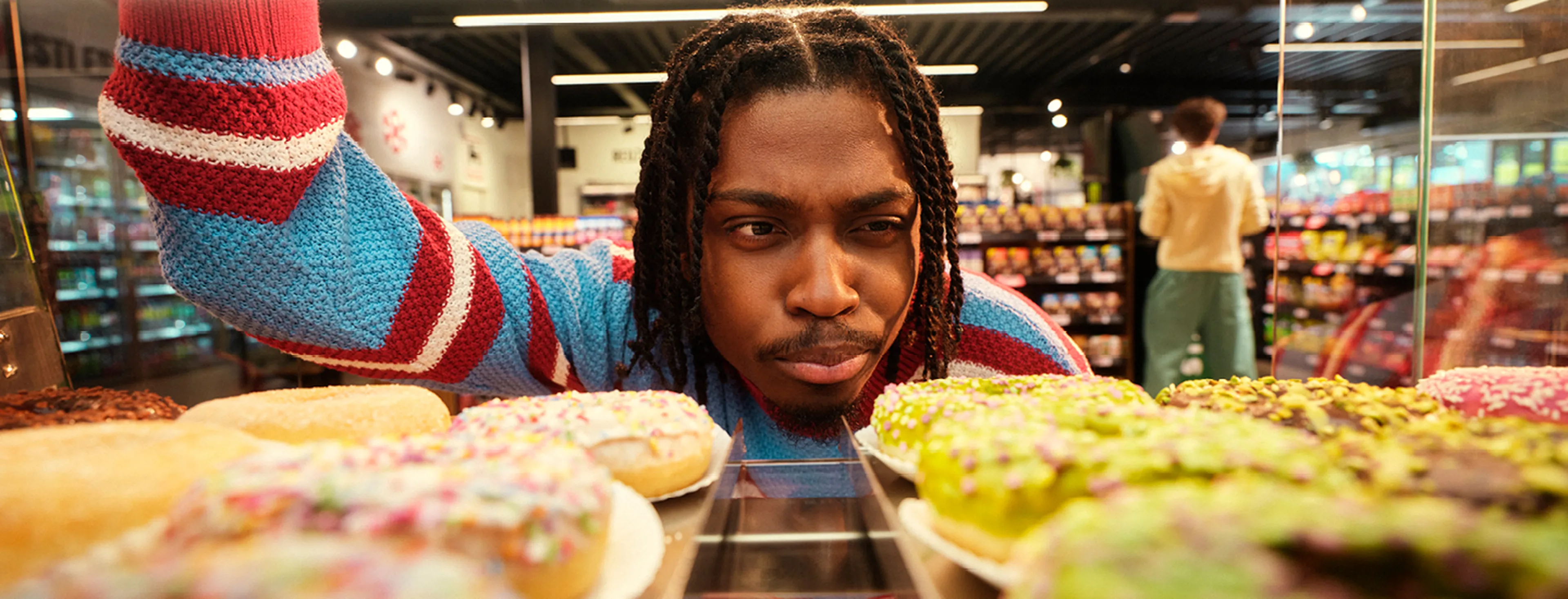 Personne avec des dreadlocks regarde des donuts en magasin ; autre personne à l’arrière-plan dans un rayon de snacks.