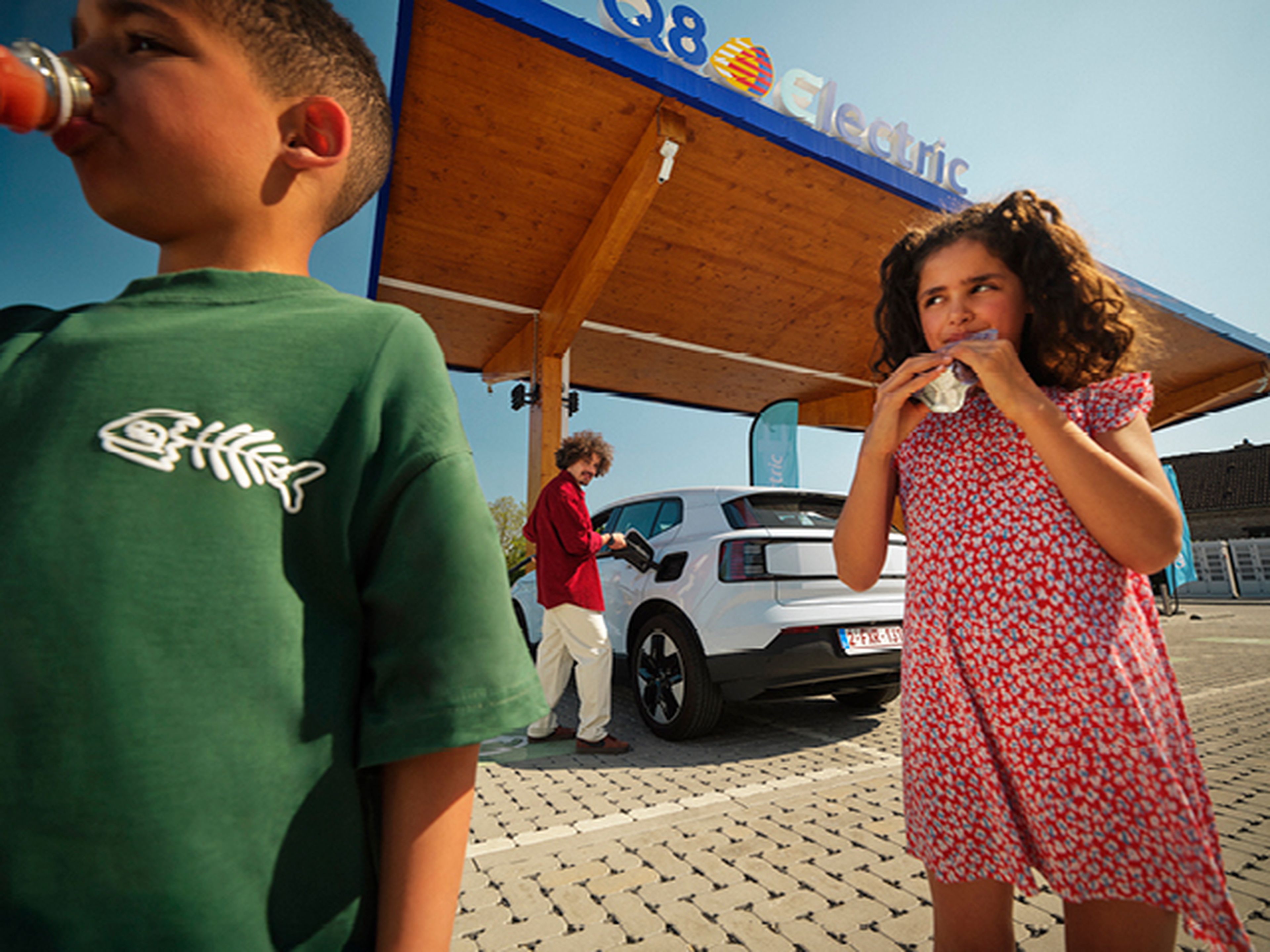Enfants boivent pendant qu'un homme recharge une voiture électrique à une station Q8 Electric.