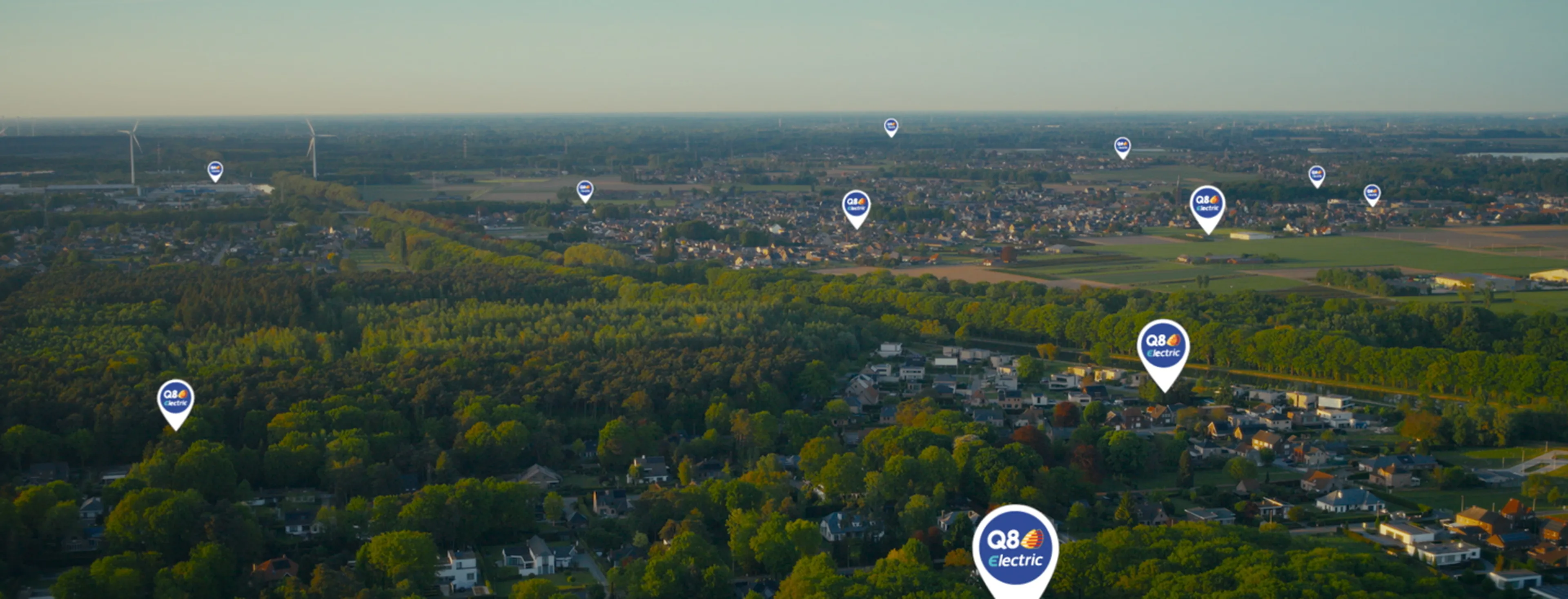 Luchtbeeld van een groene omgeving met woonwijken, windmolens en meerdere “Q8 electric”-locatiepinnen verspreid over het landschap, die laadpunten voor elektrische voertuigen aanduiden.