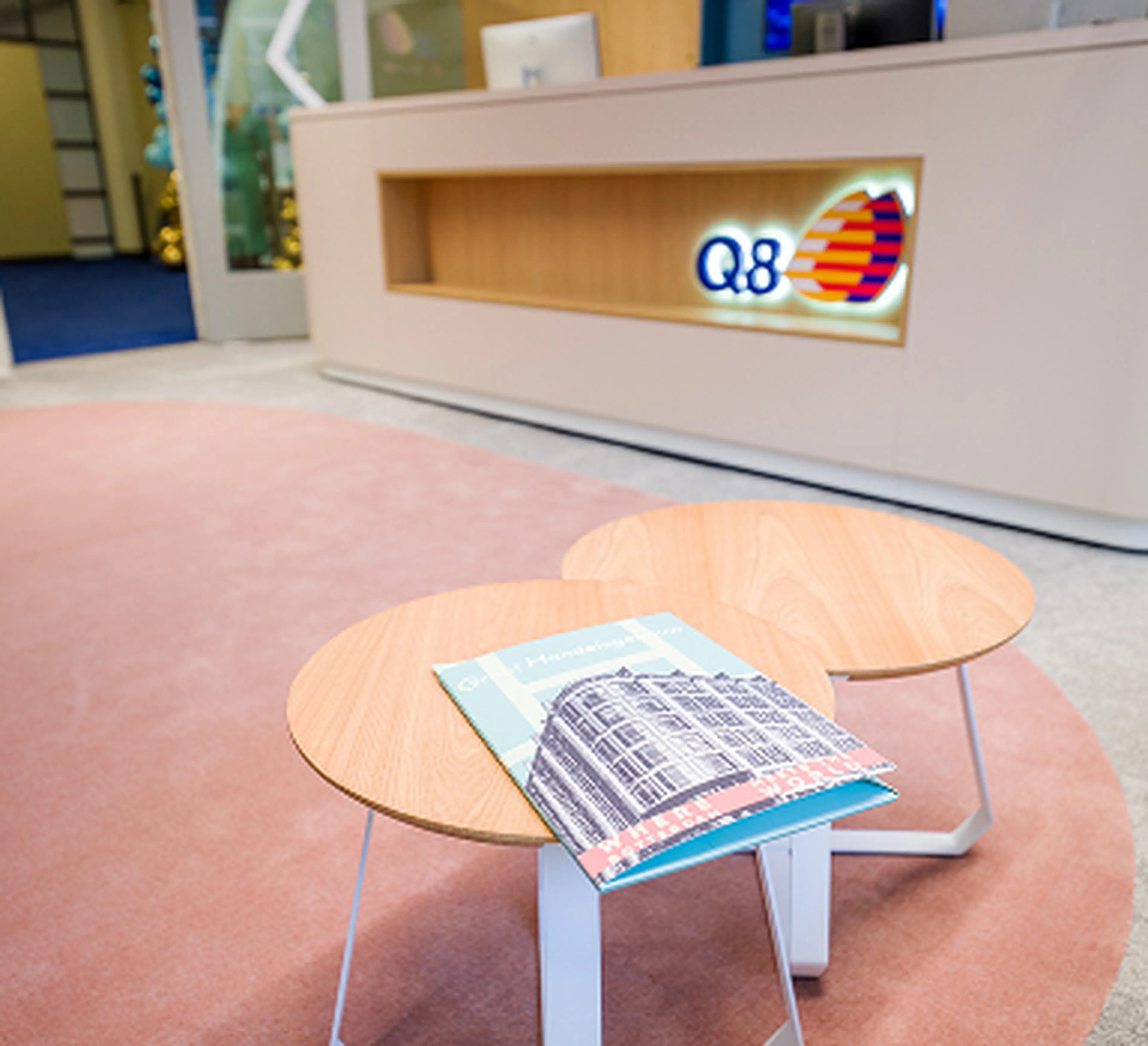 Accueil de Q8 au siège à Rotterdam, avec le logo visible sur le mur. Au premier plan, deux petites tables rondes en bois avec un livre posé dessus, sur un tapis rose pâle.
