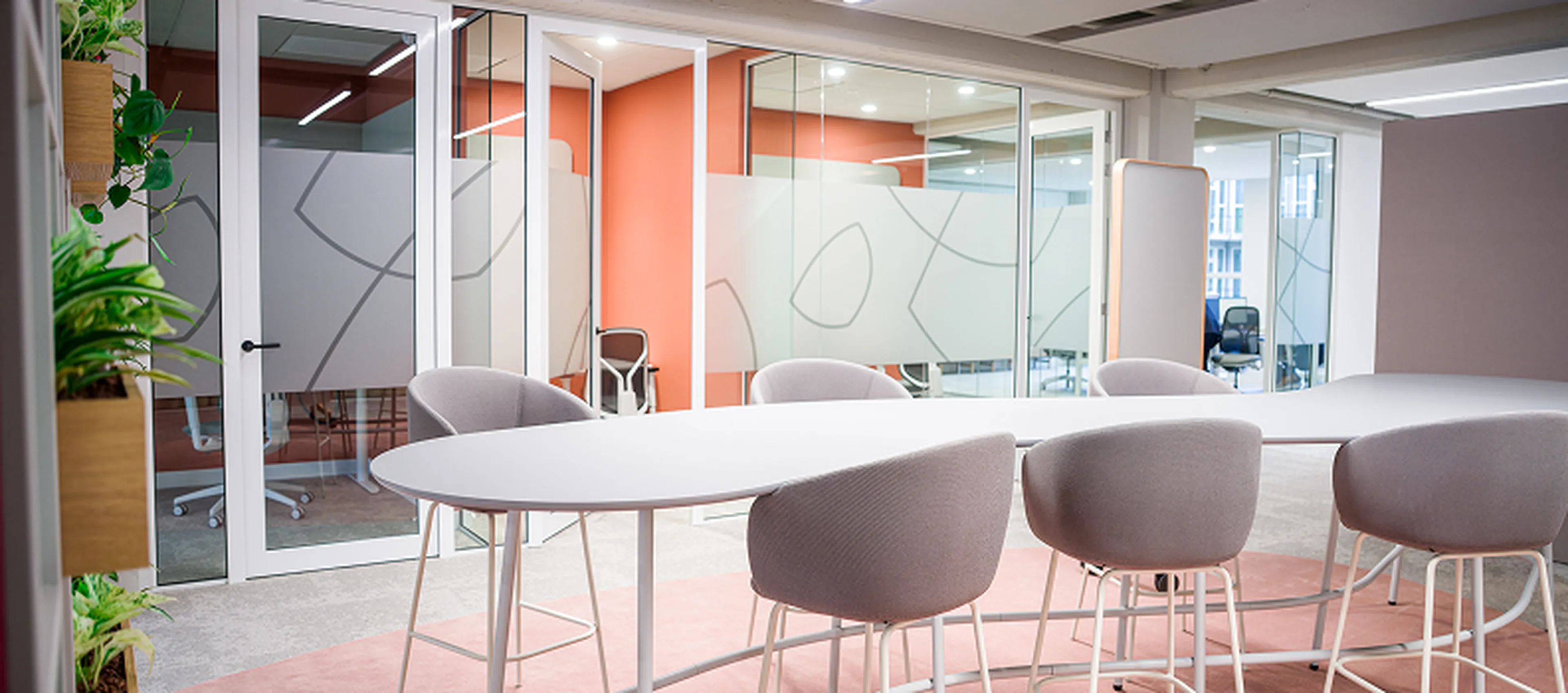 Salle de réunion au siège de Q8 à Rotterdam avec des tables rondes, des chaises hautes et beaucoup de verre. Des couleurs chaudes et des plantes créent une ambiance ouverte et fraîche.