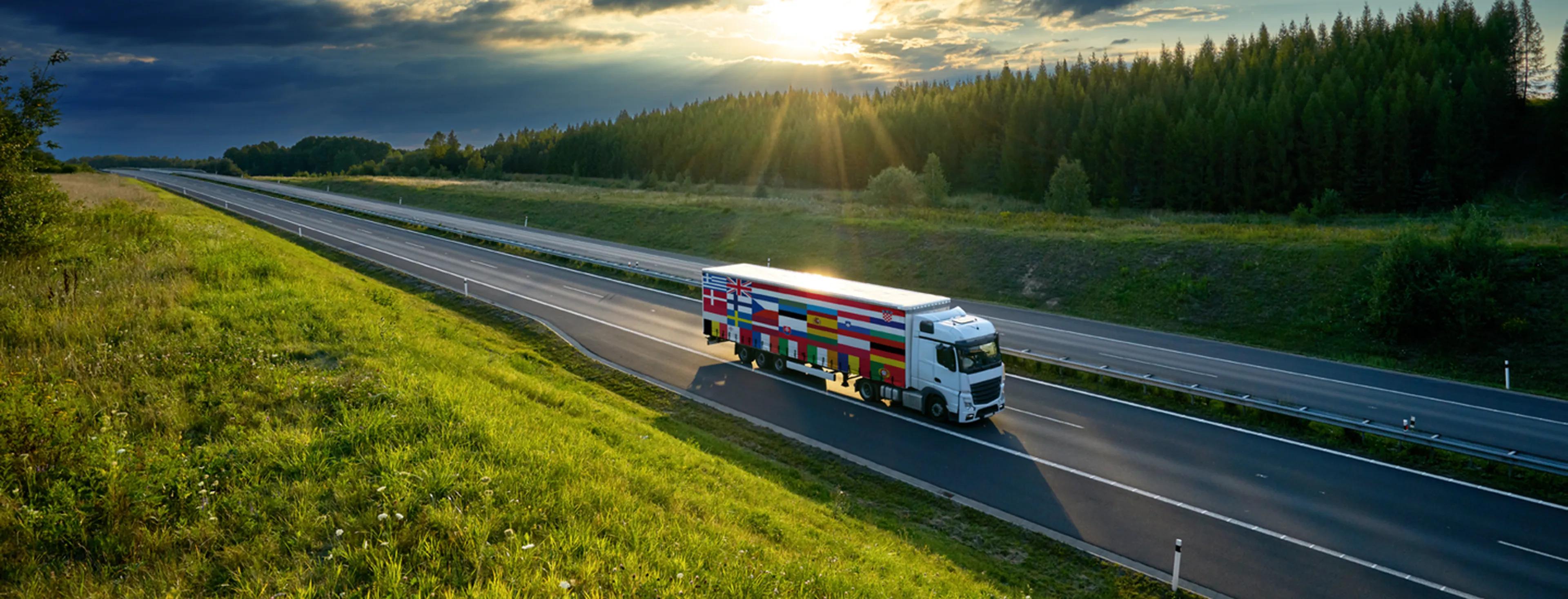 Camion avec des drapeaux européens sur le côté roulant sur une autoroute vide, entourée d’herbe verte et d’une lisière de forêt, avec des rayons de soleil perçant à travers les nuages.