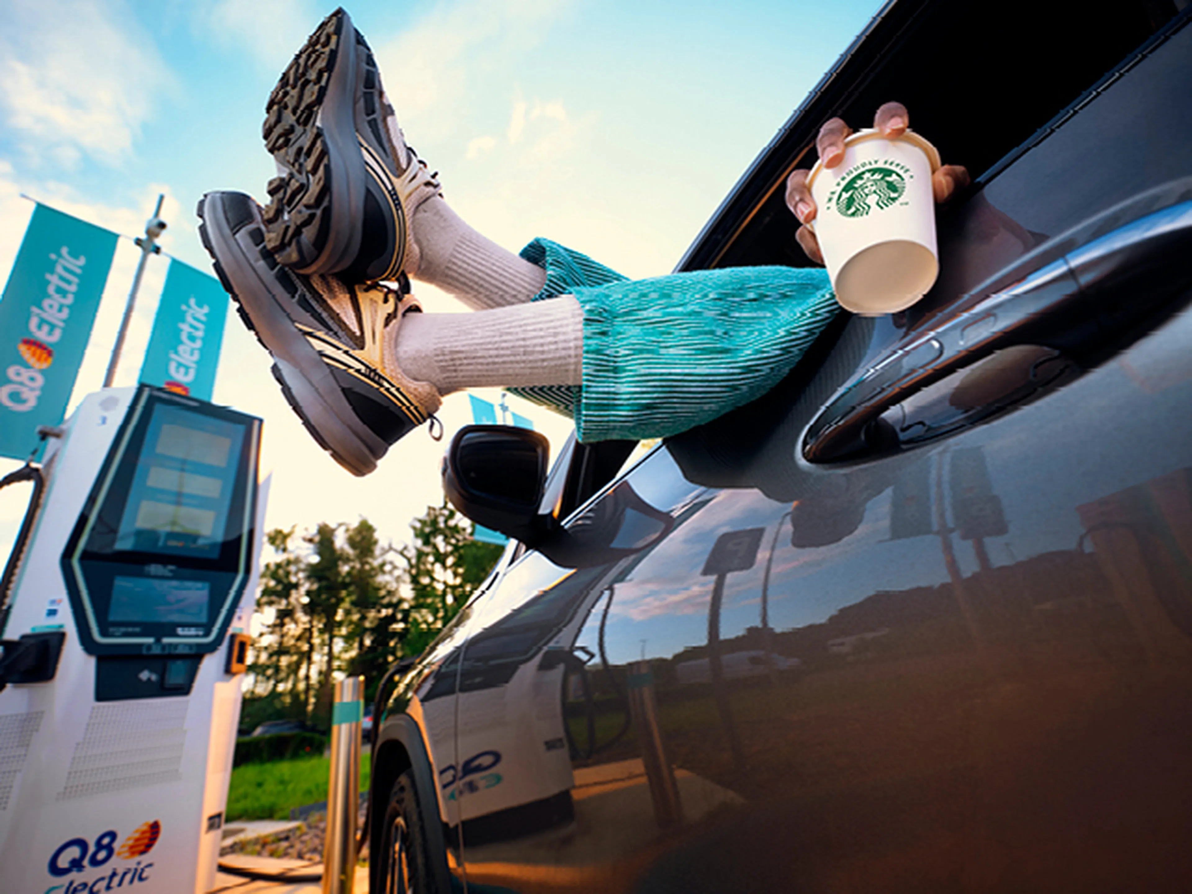 Pieds sortant de la fenêtre de voiture, personne boit du café à la station de recharge Q8 Electric.